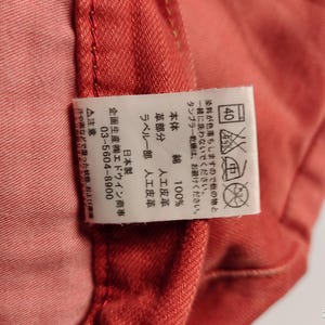 May include: Close-up of a section of red-orange fabric with a white tag. The tag has Japanese text and care symbols, including a 40-degree wash symbol. The fabric appears to be cotton, with a leather-like detail.