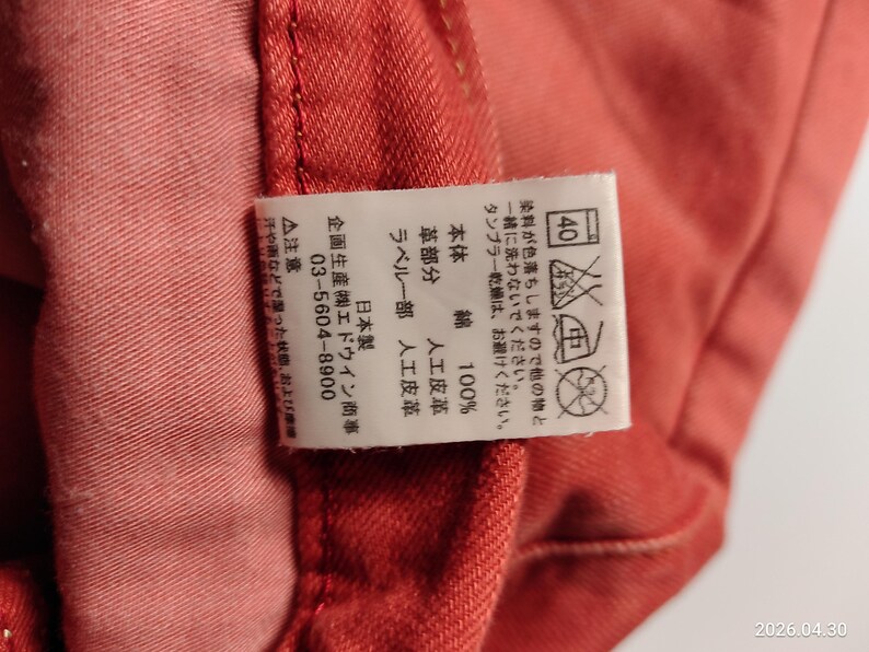 May include: Close-up of a section of red-orange fabric with a white tag. The tag has Japanese text and care symbols, including a 40-degree wash symbol. The fabric appears to be cotton, with a leather-like detail.