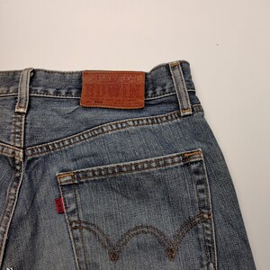 May include: Close-up of the back of blue denim jeans. The jeans have a brown leather patch with the brand name "EDWIN" and a red tag on the back pocket. The stitching is visible in a contrasting brown color.