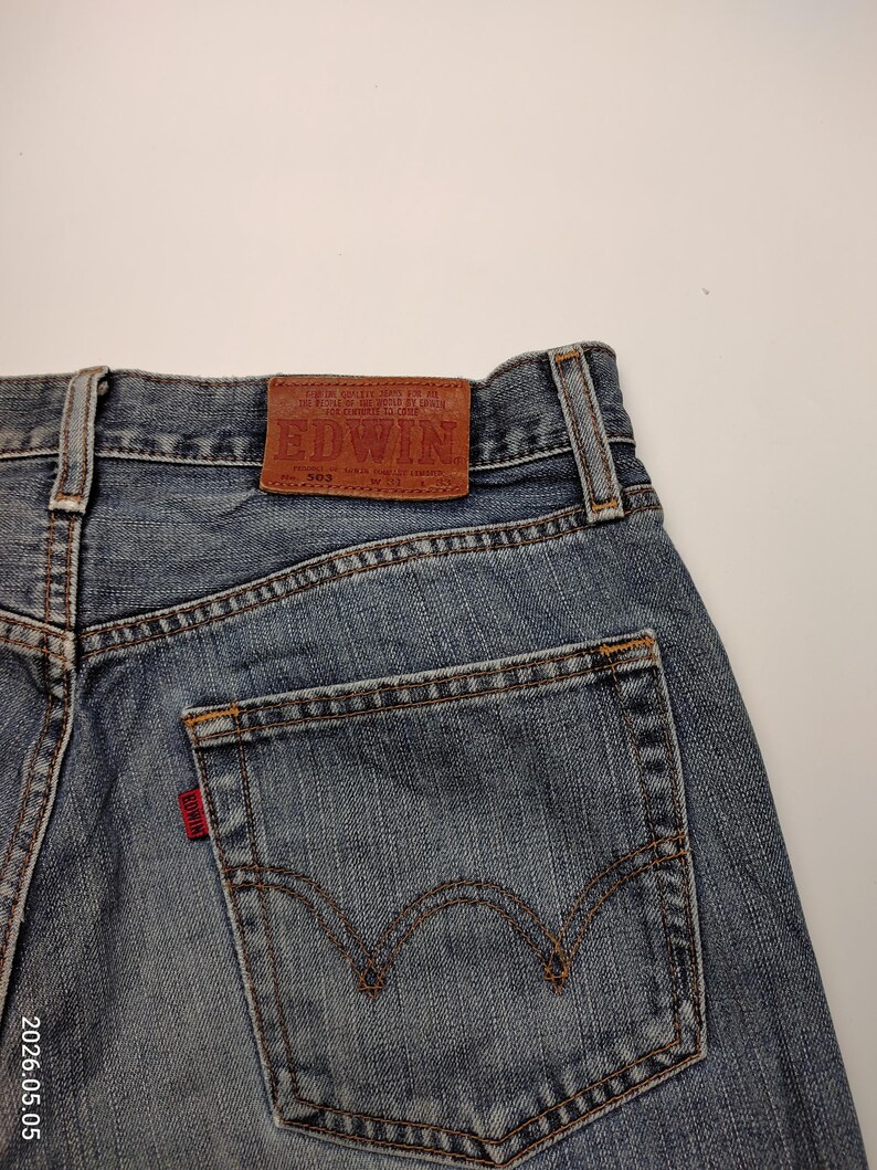 May include: Close-up of the back of blue denim jeans. The jeans have a brown leather patch with the brand name "EDWIN" and a red tag on the back pocket. The stitching is visible in a contrasting brown color.