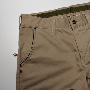 May include: Close-up of khaki-colored pants. The pants have a button closure, belt loops, and a pocket with a black trim. The stitching is in a contrasting yellow color. The waistband has a green and brown striped lining.