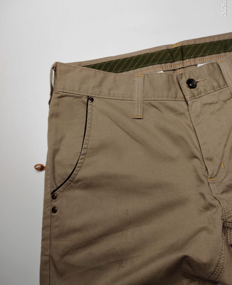 May include: Close-up of khaki-colored pants. The pants have a button closure, belt loops, and a pocket with a black trim. The stitching is in a contrasting yellow color. The waistband has a green and brown striped lining.