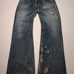 Peut inclure: Jean bleu d&eacute;lav&eacute; avec des d&eacute;tails us&eacute;s, notamment des d&eacute;chirures et des patchs us&eacute;s. Le jean a une conception classique &agrave; cinq poches et une coupe d&eacute;contract&eacute;e. Le denim montre des signes d'usure et de d&eacute;coloration.