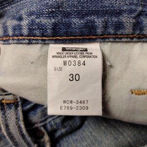 May include: Close-up of a blue denim jeans label. The label reads "Wrangler" and includes the text "MADE UNDER LICENSE FROM WRANGLER APPAREL CORPORATION". The size is indicated as "30". Additional codes are also visible.