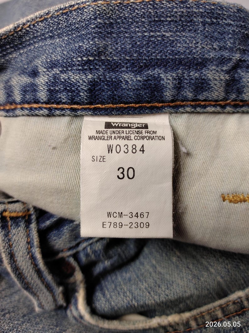 May include: Close-up of a blue denim jeans label. The label reads "Wrangler" and includes the text "MADE UNDER LICENSE FROM WRANGLER APPAREL CORPORATION". The size is indicated as "30". Additional codes are also visible.
