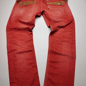 May include: Red denim jeans with brown suede pocket accents and zipper details. The jeans have a classic five-pocket design with a red Levi's tag. The back pockets feature decorative stitching and a leather patch with text.