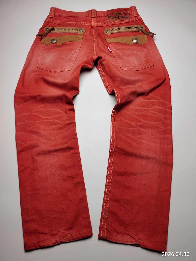 May include: Red denim jeans with brown suede pocket accents and zipper details. The jeans have a classic five-pocket design with a red Levi's tag. The back pockets feature decorative stitching and a leather patch with text.