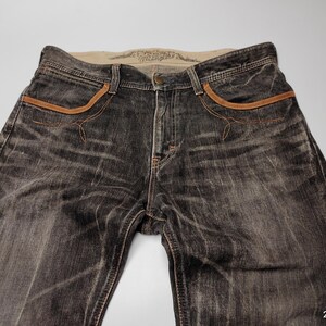 May include: Faded black denim jeans with brown pocket trim and stitching. The jeans have a classic five-pocket design and a button closure. The denim has a worn, distressed look.