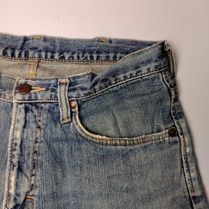 May include: Close-up of a pair of faded blue denim jeans. The jeans have a classic five-pocket design, a button closure, and a small coin pocket. The denim shows signs of wear and tear, with light fading and distressing.
