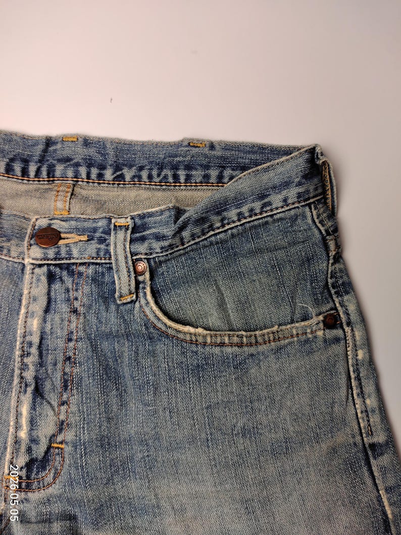 May include: Close-up of a pair of faded blue denim jeans. The jeans have a classic five-pocket design, a button closure, and a small coin pocket. The denim shows signs of wear and tear, with light fading and distressing.