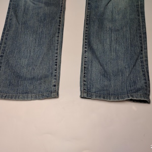 May include: Close-up of the lower portion of a pair of blue denim jeans. The jeans have a classic five-pocket design and a slightly faded wash. The hem of the jeans is visible, showing a worn edge. The jeans are laid flat on a white surface.