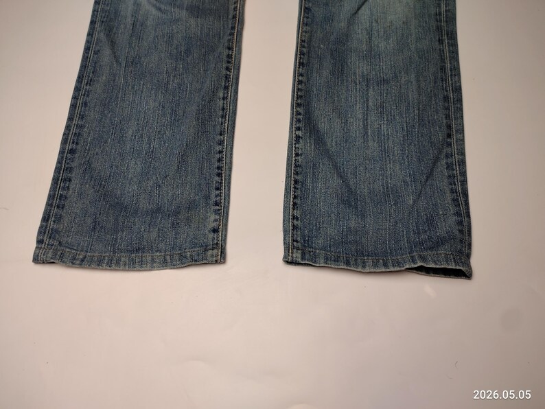 May include: Close-up of the lower portion of a pair of blue denim jeans. The jeans have a classic five-pocket design and a slightly faded wash. The hem of the jeans is visible, showing a worn edge. The jeans are laid flat on a white surface.