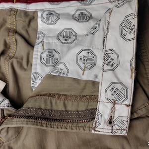 May include: Close-up of khaki-colored pants with a visible zipper and button closure. The interior lining features a repeating pattern of the brand name "EDWIN" and the text "SINCE 1961". A tag with text is visible.