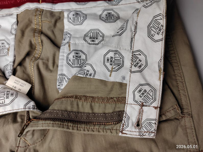 May include: Close-up of khaki-colored pants with a visible zipper and button closure. The interior lining features a repeating pattern of the brand name "EDWIN" and the text "SINCE 1961". A tag with text is visible.