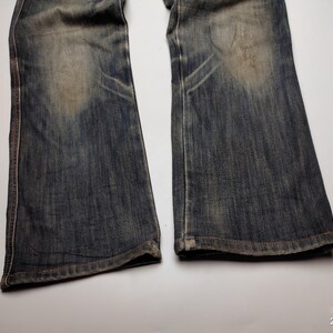 Puede incluir: Primer plano de un par de vaqueros de mezclilla azul descoloridos. Los vaqueros tienen un estilo de corte de bota con desgaste visible, incluyendo parches m&aacute;s claros y &aacute;reas desgastadas. La costura es visible a lo largo de las costuras.