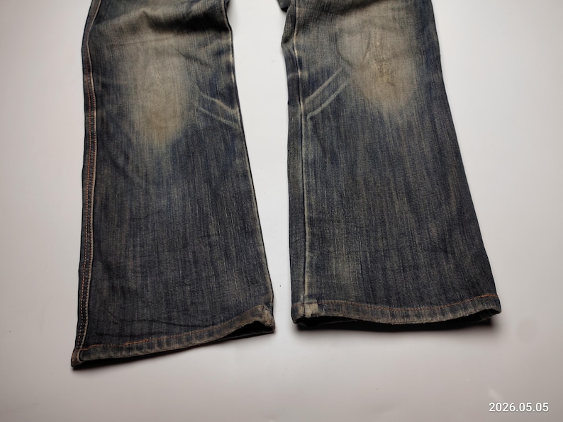 Puede incluir: Primer plano de un par de vaqueros de mezclilla azul descoloridos. Los vaqueros tienen un estilo de corte de bota con desgaste visible, incluyendo parches m&aacute;s claros y &aacute;reas desgastadas. La costura es visible a lo largo de las costuras.