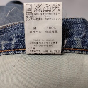 May include: Close-up of a denim garment with a white tag. The tag displays washing instructions, fabric composition (100% cotton), and a "Made in China" label. The tag also includes Japanese text and a phone number.