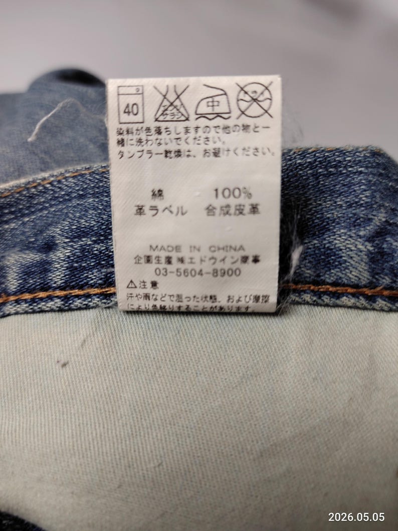 May include: Close-up of a denim garment with a white tag. The tag displays washing instructions, fabric composition (100% cotton), and a "Made in China" label. The tag also includes Japanese text and a phone number.