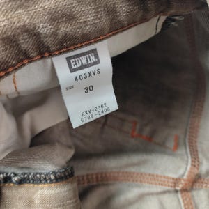 May include: Close-up of brown denim jeans with orange stitching. The tag reads "EDWIN" and "403XVS", with a size of 30. The jeans have a faded appearance and visible seams.