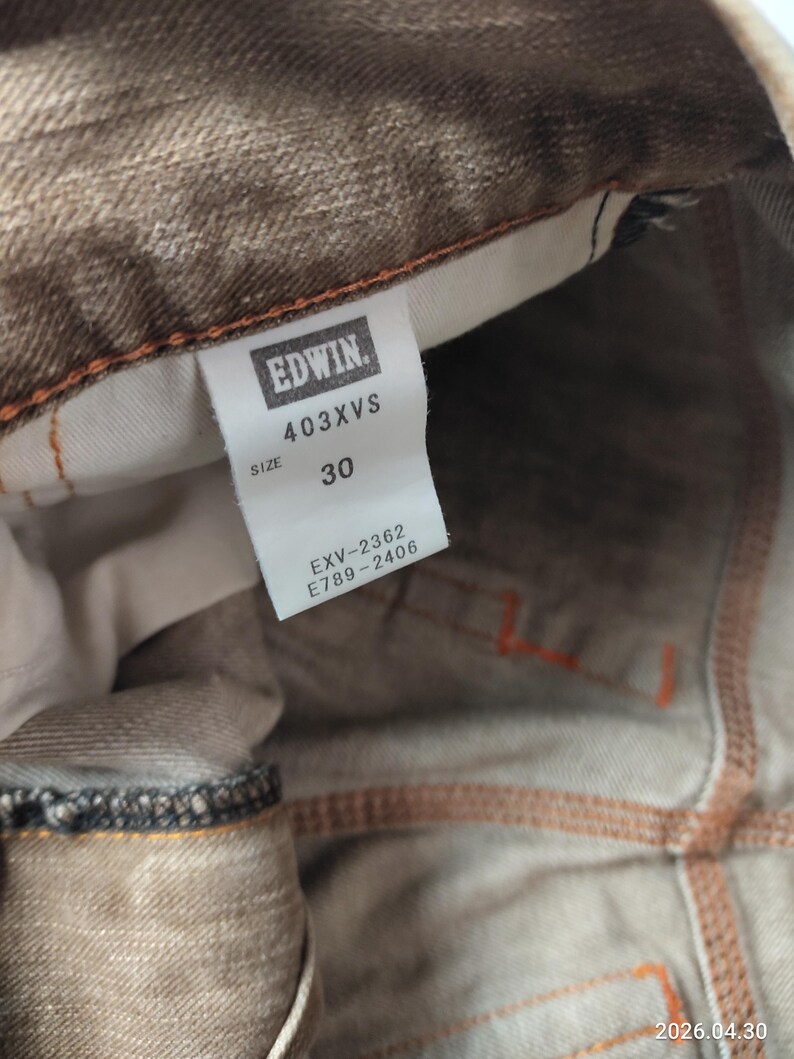 May include: Close-up of brown denim jeans with orange stitching. The tag reads "EDWIN" and "403XVS", with a size of 30. The jeans have a faded appearance and visible seams.