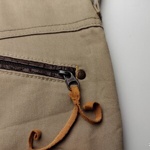 May include: Close-up of tan-colored pants with a brown leather-trimmed pocket and a black zipper. The zipper pull is a tan leather cord. A small, round, bronze-colored button is visible.