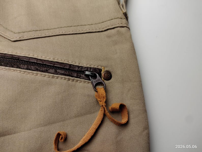 May include: Close-up of tan-colored pants with a brown leather-trimmed pocket and a black zipper. The zipper pull is a tan leather cord. A small, round, bronze-colored button is visible.
