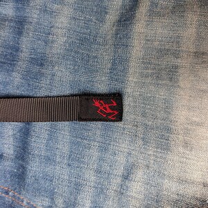 May include: A close-up of a black woven strap with a red logo on a black label. The strap is set against a blue denim background. The logo is a stylized figure. The denim has a faded, worn appearance.
