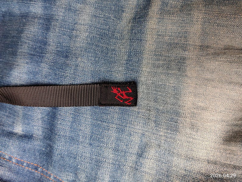 May include: A close-up of a black woven strap with a red logo on a black label. The strap is set against a blue denim background. The logo is a stylized figure. The denim has a faded, worn appearance.