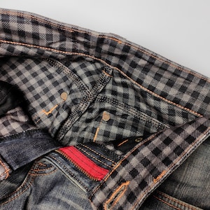 Puede incluir: Primer plano de unos vaqueros de mezclilla con un forro a cuadros en la cintura y los bolsillos. Los vaqueros tienen una cremallera roja y costuras naranjas. La tela parece desgastada y descolorida, con un dise&ntilde;o cl&aacute;sico de cinco bolsillos.