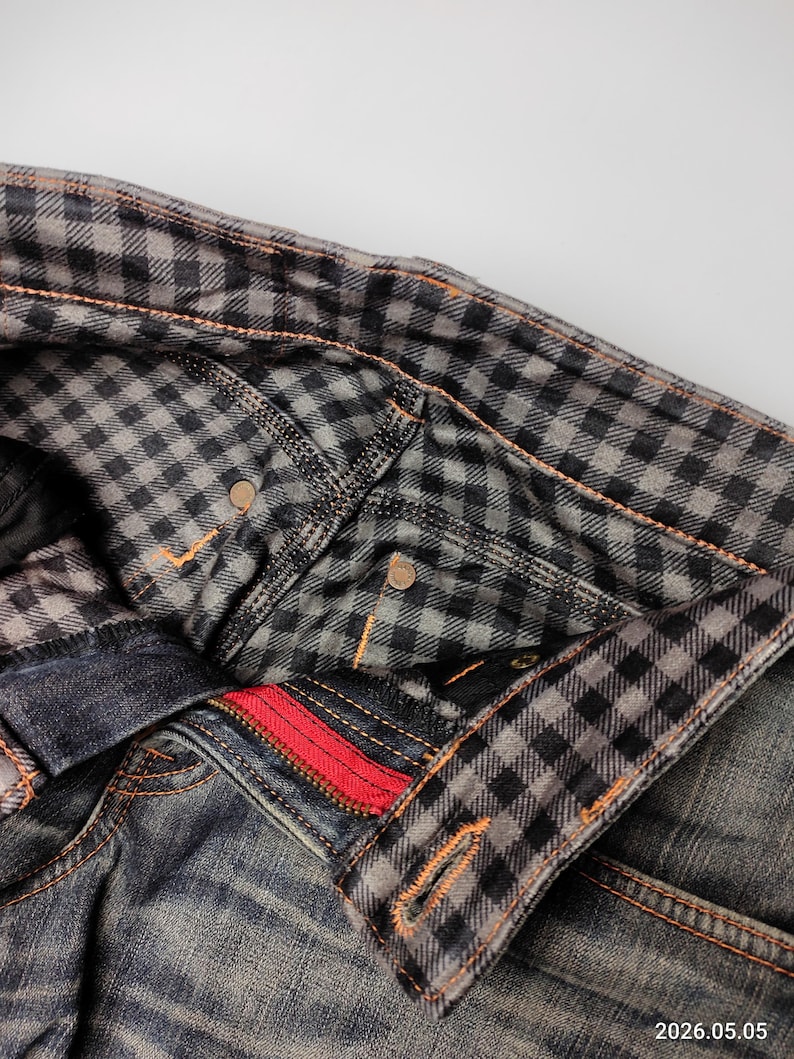 Puede incluir: Primer plano de unos vaqueros de mezclilla con un forro a cuadros en la cintura y los bolsillos. Los vaqueros tienen una cremallera roja y costuras naranjas. La tela parece desgastada y descolorida, con un dise&ntilde;o cl&aacute;sico de cinco bolsillos.