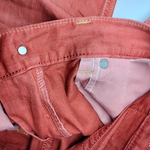 May include: Close-up of a pair of red-orange denim jeans. The image highlights the waistband, button, and stitching details. The interior pocket lining is a lighter shade of pink.