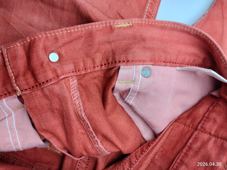 May include: Close-up of a pair of red-orange denim jeans. The image highlights the waistband, button, and stitching details. The interior pocket lining is a lighter shade of pink.