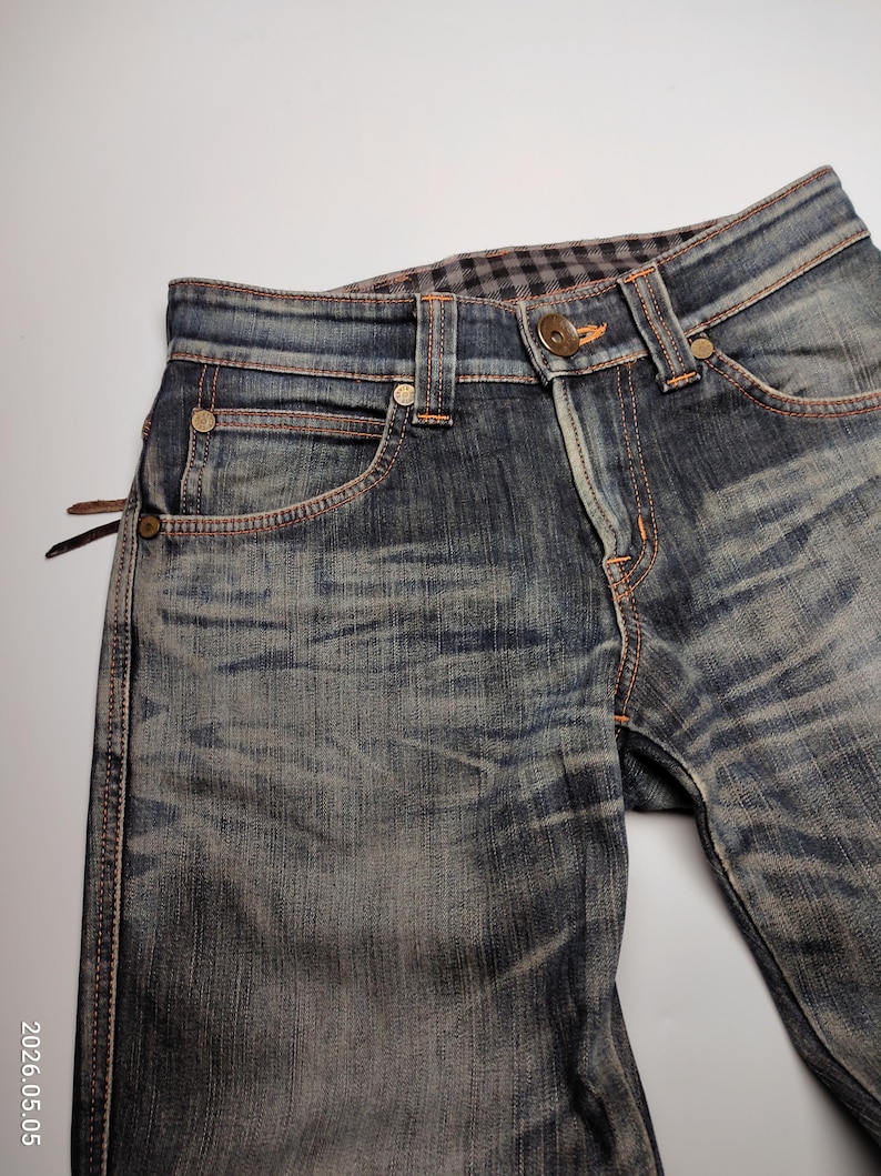 Puede incluir: Primer plano de unos vaqueros de mezclilla azul con un lavado descolorido y costuras naranjas. Los vaqueros tienen remaches met&aacute;licos y un cierre de bot&oacute;n en la cintura, que tiene un forro a cuadros. El dise&ntilde;o incluye un dise&ntilde;o cl&aacute;sico de cinco bolsillos.