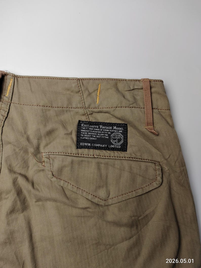 May include: Close-up of khaki-colored pants with a back pocket and a label that reads "EXCLUSIVE VINTAGE MODEL" and "EDWIN COMPANY LIMITED". The pants have visible stitching and a brown belt loop. The date "2026.05.01" is in the bottom right corner.