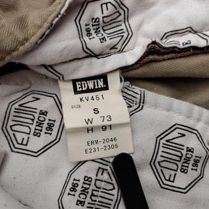 May include: Close-up of the inside of a pair of khaki-colored pants. A white tag with the brand name "EDWIN" and size information is visible. The tag reads "W 73" and "H 91". The lining features a repeating black and white logo.