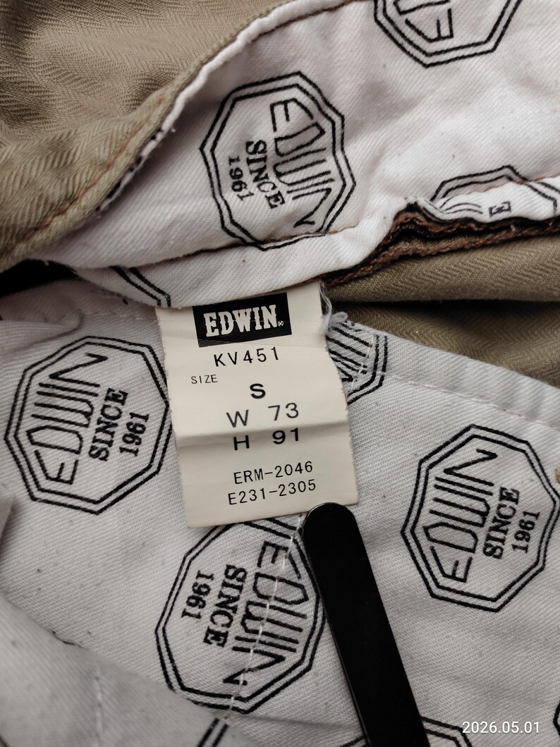 May include: Close-up of the inside of a pair of khaki-colored pants. A white tag with the brand name "EDWIN" and size information is visible. The tag reads "W 73" and "H 91". The lining features a repeating black and white logo.