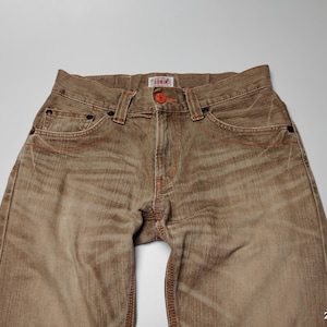 May include: Close-up of brown denim jeans with red stitching details. The jeans have a button closure and pockets. The brand name "Edwin" is visible on a small tag. The jeans are a neutral color and appear to be pre-worn.