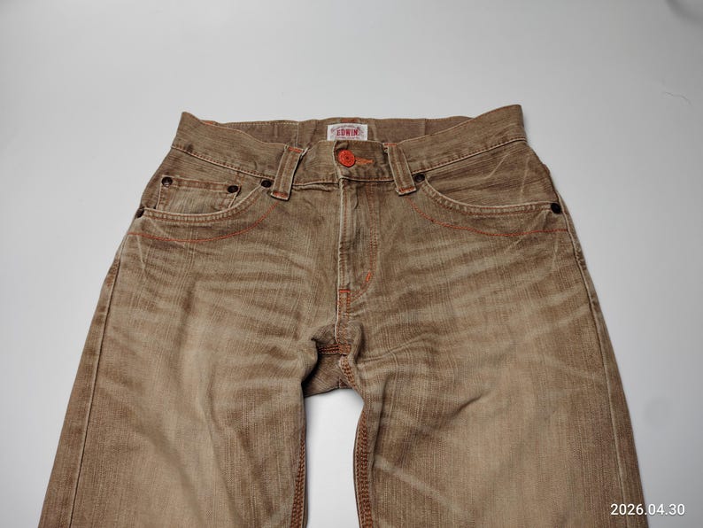 May include: Close-up of brown denim jeans with red stitching details. The jeans have a button closure and pockets. The brand name "Edwin" is visible on a small tag. The jeans are a neutral color and appear to be pre-worn.