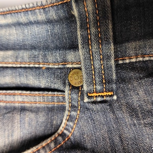 Puede incluir: Primer plano de unos vaqueros azules con costuras naranjas. Los vaqueros tienen un bolsillo peque&ntilde;o, un bot&oacute;n y una trabilla. La tela vaquera tiene un aspecto descolorido.
