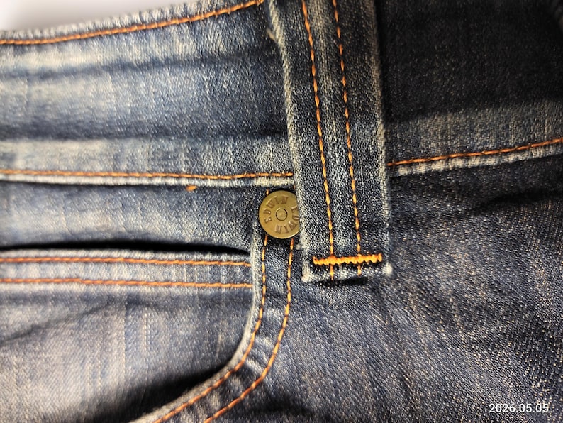Puede incluir: Primer plano de unos vaqueros azules con costuras naranjas. Los vaqueros tienen un bolsillo peque&ntilde;o, un bot&oacute;n y una trabilla. La tela vaquera tiene un aspecto descolorido.