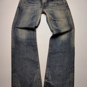 May include: Faded blue denim jeans with a classic five-pocket design. The jeans have a straight leg cut and show signs of wear, with lighter patches and subtle distressing. The stitching is a contrasting orange-brown color.