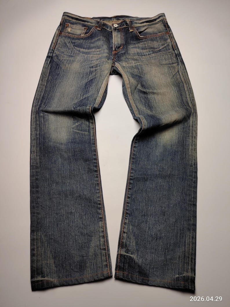 May include: Faded blue denim jeans with a classic five-pocket design. The jeans have a straight leg cut and show signs of wear, with lighter patches and subtle distressing. The stitching is a contrasting orange-brown color.