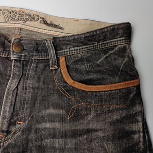 May include: Close-up of a pair of dark gray Wrangler jeans. The jeans feature a brown leather-trimmed pocket and a button closure. The Wrangler logo is visible on the waistband. The jeans have a worn, distressed look.