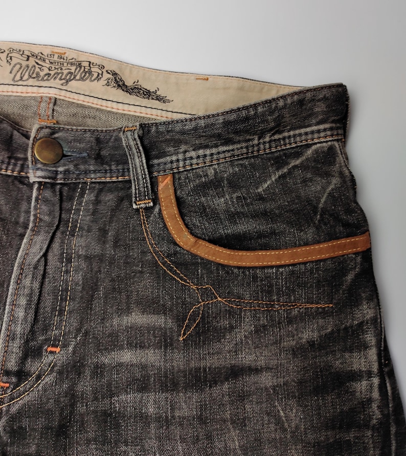 May include: Close-up of a pair of dark gray Wrangler jeans. The jeans feature a brown leather-trimmed pocket and a button closure. The Wrangler logo is visible on the waistband. The jeans have a worn, distressed look.