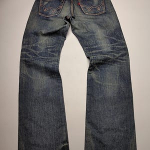 May include: Faded blue denim jeans with classic five-pocket design. Features red stitching, a red logo tag, and a worn appearance. The jeans are laid flat, showcasing the back pockets and the overall distressed look.