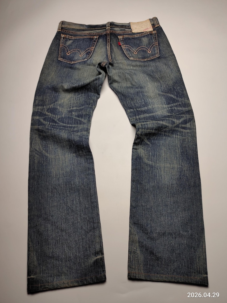 May include: Faded blue denim jeans with classic five-pocket design. Features red stitching, a red logo tag, and a worn appearance. The jeans are laid flat, showcasing the back pockets and the overall distressed look.
