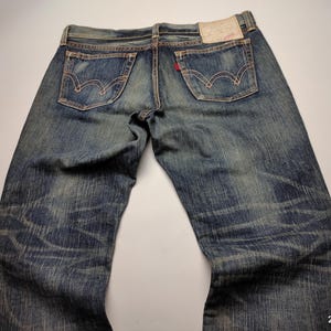 May include: Faded blue denim jeans with classic five-pocket styling. The jeans feature a worn appearance with visible fading and distressing. The back pockets have a distinctive curved stitch design. A red tag is visible on the back pocket.