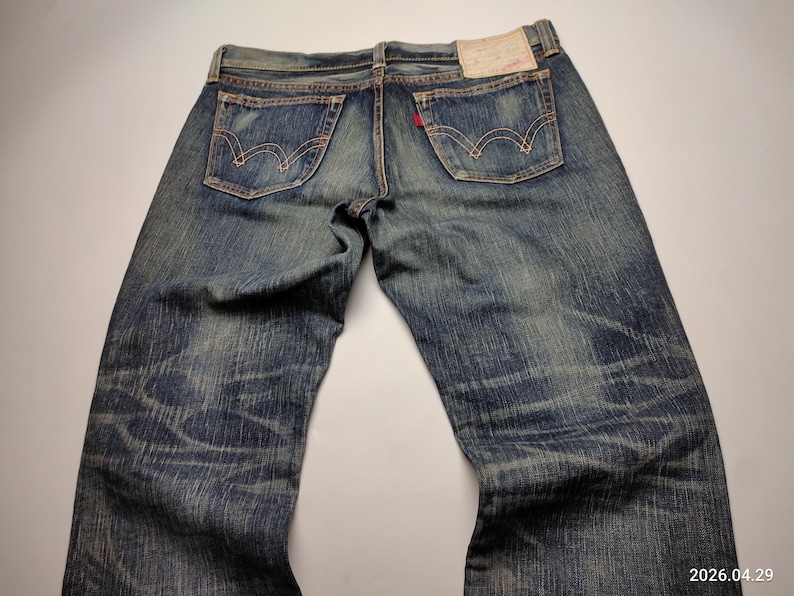 May include: Faded blue denim jeans with classic five-pocket styling. The jeans feature a worn appearance with visible fading and distressing. The back pockets have a distinctive curved stitch design. A red tag is visible on the back pocket.