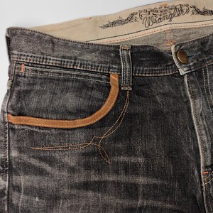 May include: Close-up of dark gray denim jeans with tan pocket trim and stitching. The jeans feature the "Wrangler" logo and text on the waistband. The jeans have a button closure and a classic five-pocket design.