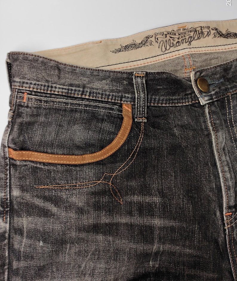 May include: Close-up of dark gray denim jeans with tan pocket trim and stitching. The jeans feature the "Wrangler" logo and text on the waistband. The jeans have a button closure and a classic five-pocket design.